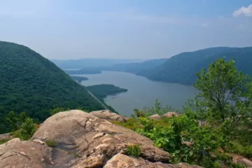 Scenic overlook on a rocky mountain, with forest mountains and a large river before a clear sky.